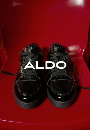 Schwarze Sneakers mit einer strukturierten Oberfläche, glänzender Optik und schwarzen Schnürsenkeln. Markenname "ALDO" auf der Zunge gedruckt. Vor einem roten Hintergrund.