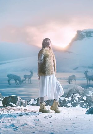 Modèle portant un gilet en fourrure léger et duveteux par-dessus une robe blanche, chaussé de bottes d'hiver beiges, se tenant sur des rochers enneigés, avec des rennes en arrière-plan.