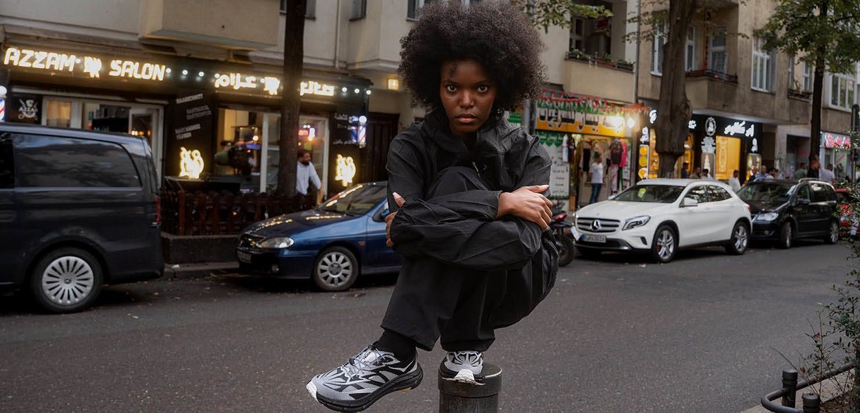 Black zip-up jacket, black pants, and patterned athletic shoes. Model squats on a pole against a city street backdrop with shops and cars.