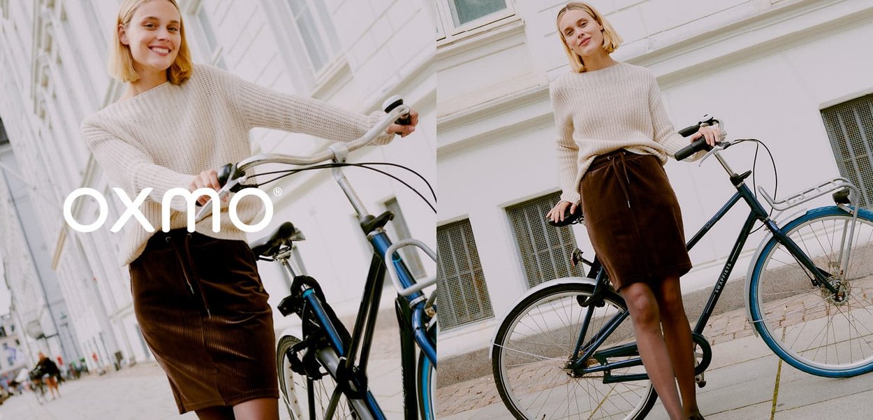 Pull en crème tricoté associé à une jupe en velours côtelé marron, se tenant à côté d'un vélo bleu sur une rue bordée de bâtiments blancs.
