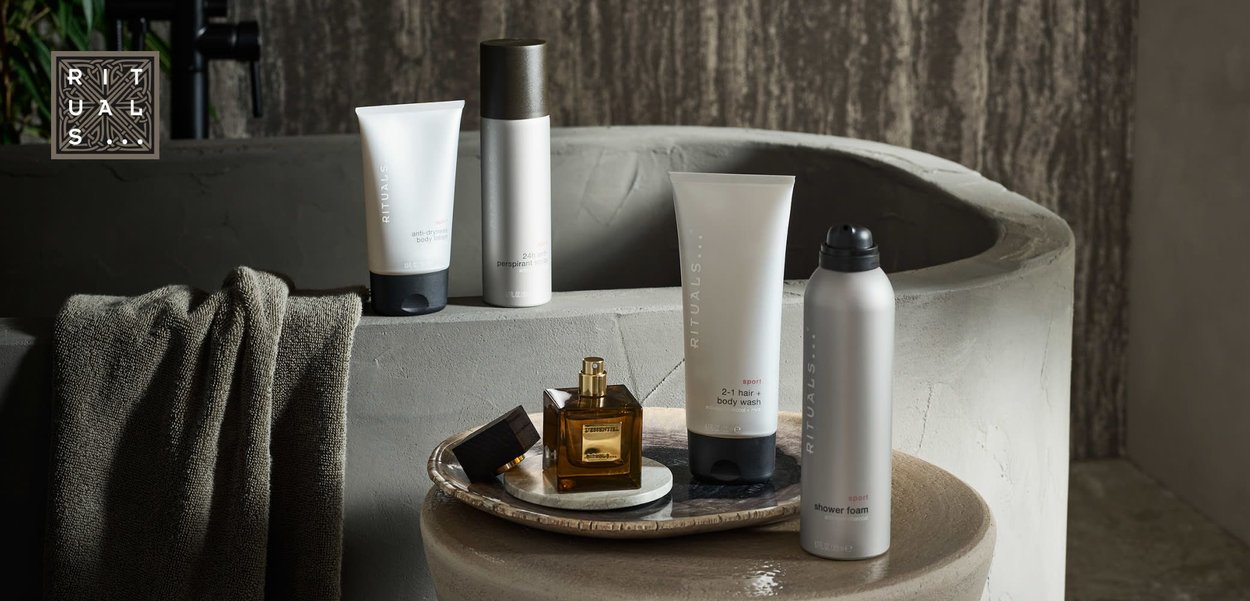Set of personal care products: white tubes with gray accents, silver spray can, gold perfume bottle on a textured tray. Concrete basin in background.