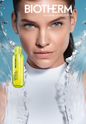 Nahaufnahme einer Frau mit klarer Haut und blauen Augen, Wasserspritzern und einer gelben Biotherm-Hautpflegeproduktflasche vor einem hellblauen Hintergrund.