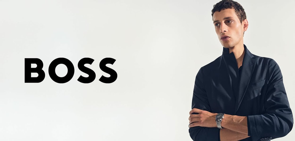 Mann im dunklen Blazer mit verschränkten Armen und Armbanduhr, steht neben großem, fettem schwarzem Text "BOSS" auf weißem Hintergrund.