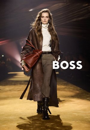 Vrouwelijk model loopt over de catwalk met een lange bruine leren jas, witte coltrui, grijze broek, zwarte laarzen, en draagt een grote bruine handtas.