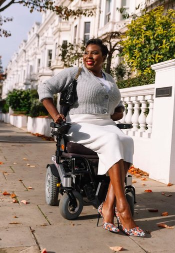Elektrisk rullstol med en grå kudde; en kvinna bär en grå kofta, vit kjol och blommiga högklackade skor, leende utomhus.