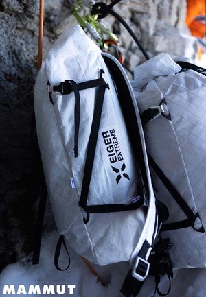 Des sacs à dos Eiger Extreme blancs avec des sangles noires suspendus près de rochers et de neige, avec du matériel d'escalade visible en arrière-plan.