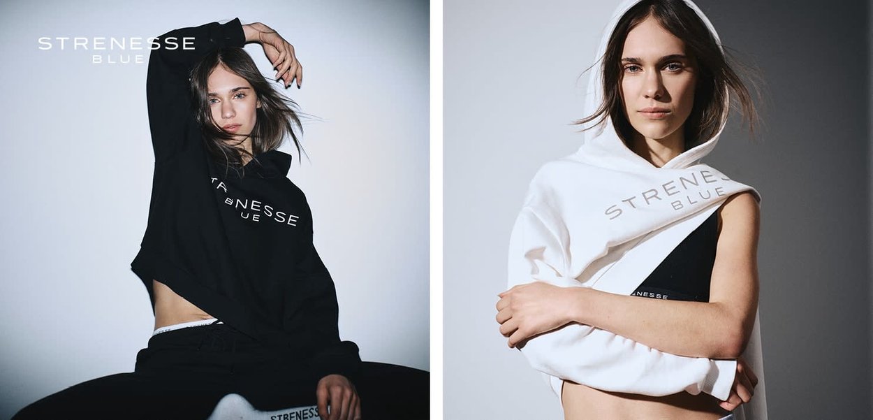 Frau präsentiert schwarze und weiße Strenesse Blue Hoodies, posiert selbstbewusst vor einfarbigem Hintergrund, mit sichtbarem Markennamen auf der Kleidung.