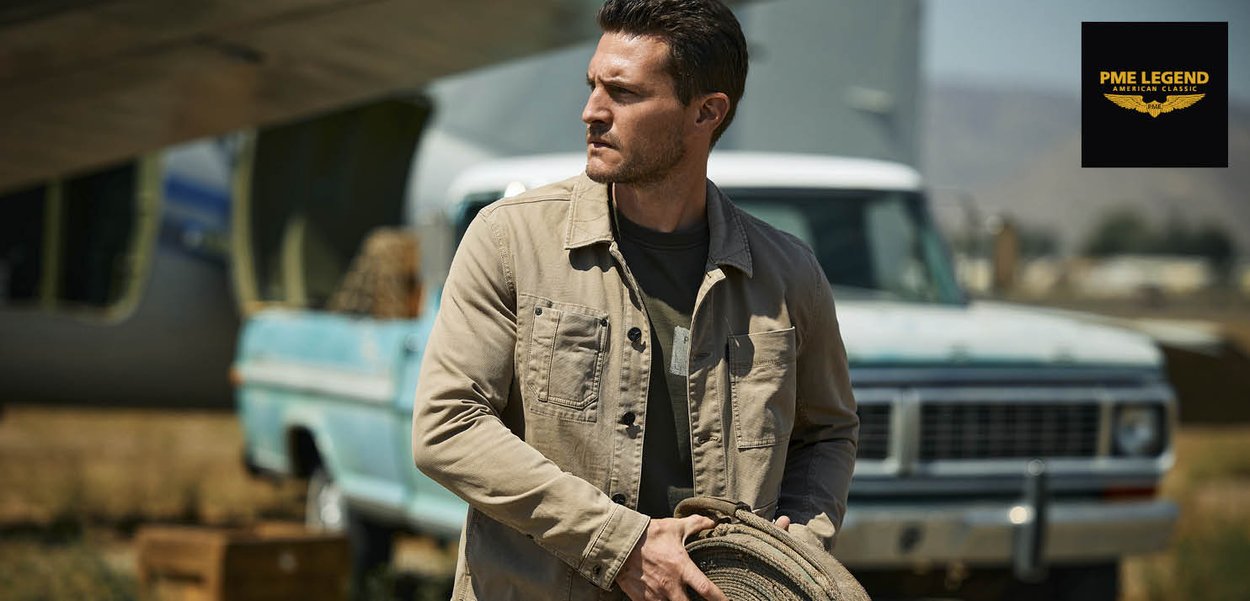 Hombre con chaqueta beige sostiene una cuerda enrollada de pie al aire libre cerca de una camioneta azul vintage bajo un puente.