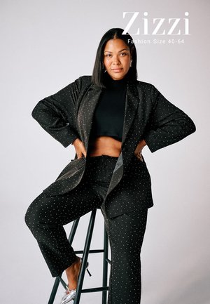 Femme portant un costume noir à pois et un crop top, assise avec confiance sur un tabouret devant un fond uni avec le texte "Zizzi Fashion Taille 40-64".
