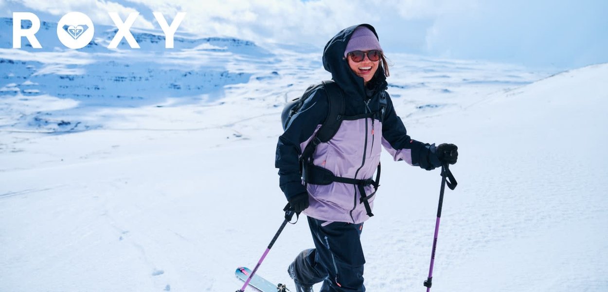 Veste de ski violette et noire avec capuche, portée avec des bâtons de ski. Paysage enneigé en arrière-plan, illustrant une activité de sports d'hiver.