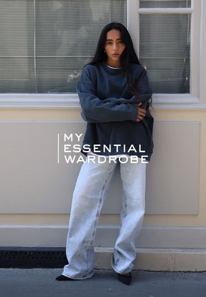 Svetlgray Oversized-Sweatshirt, hellblaue High-Waist-Jeans, schwarze spitze Schuhe. Das Modell lehnt gegen eine Wand, natürlichem Licht von einem Fenster.