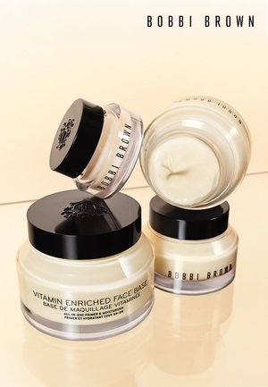 Trois pots de Base Enrichie en Vitamines de Bobbi Brown avec des couvercles noirs brillants, un contenu de couleur crème et des bases transparentes disposés de manière créative.