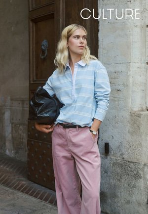 Hellblau gestreiftes Langarm-Poloshirt, weißer Kragen, rosa weit geschnittene Hose, schwarze strukturierte Handtasche, steht vor einer Steinmauer.