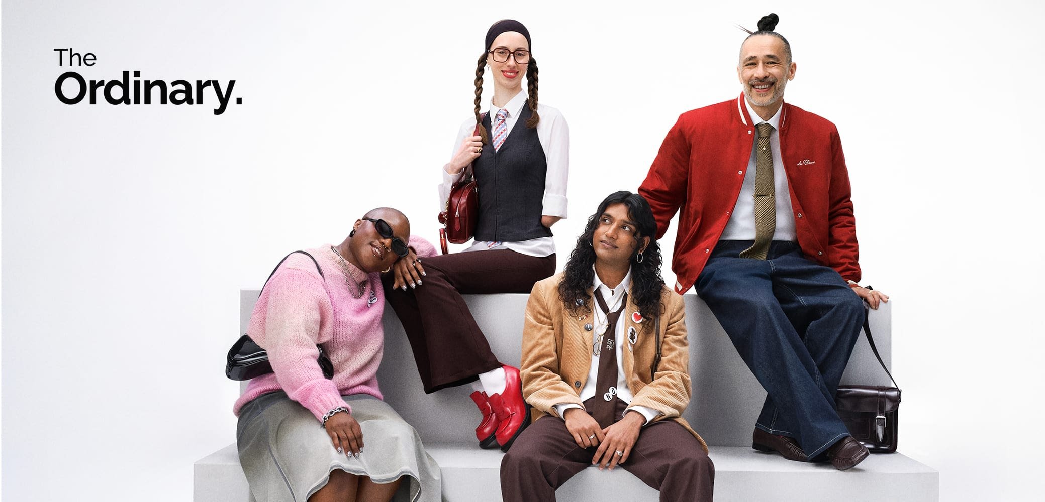 Four individuals pose on a light gray platform. They wear diverse outfits, including pastel knits, elongated jackets, and bold footwear.