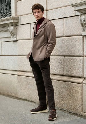 Braune Kapuzenjacke über einem burgunderfarbenen Rollkragenpullover, kombiniert mit dunklen Hosen und braunen Turnschuhen auf einem strukturierten Stein-Hintergrund.