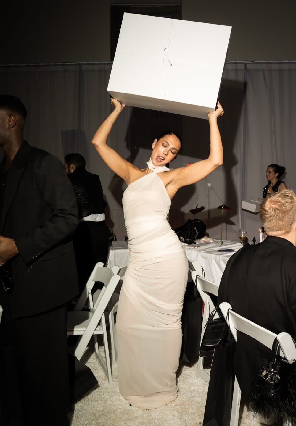 En kvinna i en figursydd, draperad ljusbeige klänning håller en stor vit låda ovanför huvudet. I bakgrunden syns sittande gäster och ett svagt upplyst rum.