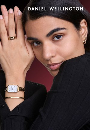 Złoty zegarek Daniel Wellington z białą kwadratową tarczą, czarnymi akcentami i metalową bransoletą, zestawiony ze złotą bransoletką typu mankiet. Ciemne tło.