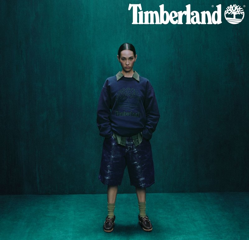 Sweter Timberland w kolorze granatowym z teksturowanym logo, w zestawieniu z ciemnymi, luźnymi szortami i zieloną koszulą w kratę. Brązowe buty dopełniają stylizację.