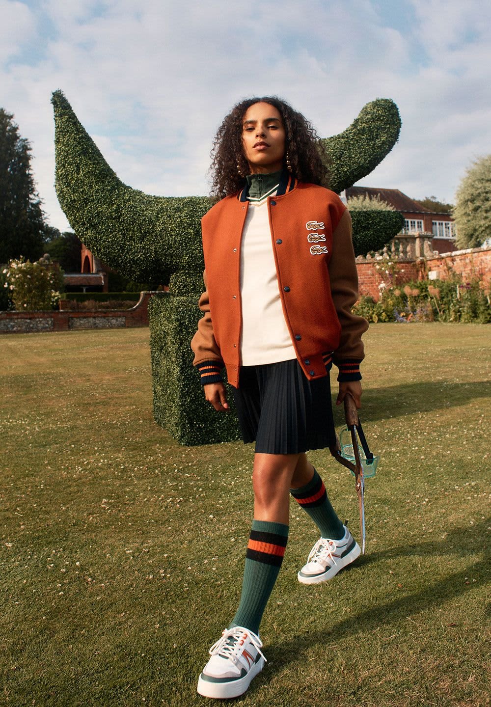 Veste de sport orange et marron, chemise blanche, jupe plissée noire, chaussettes vertes avec des rayures rouges et jaunes, chaussures de sport blanches, tenant un parapluie.
