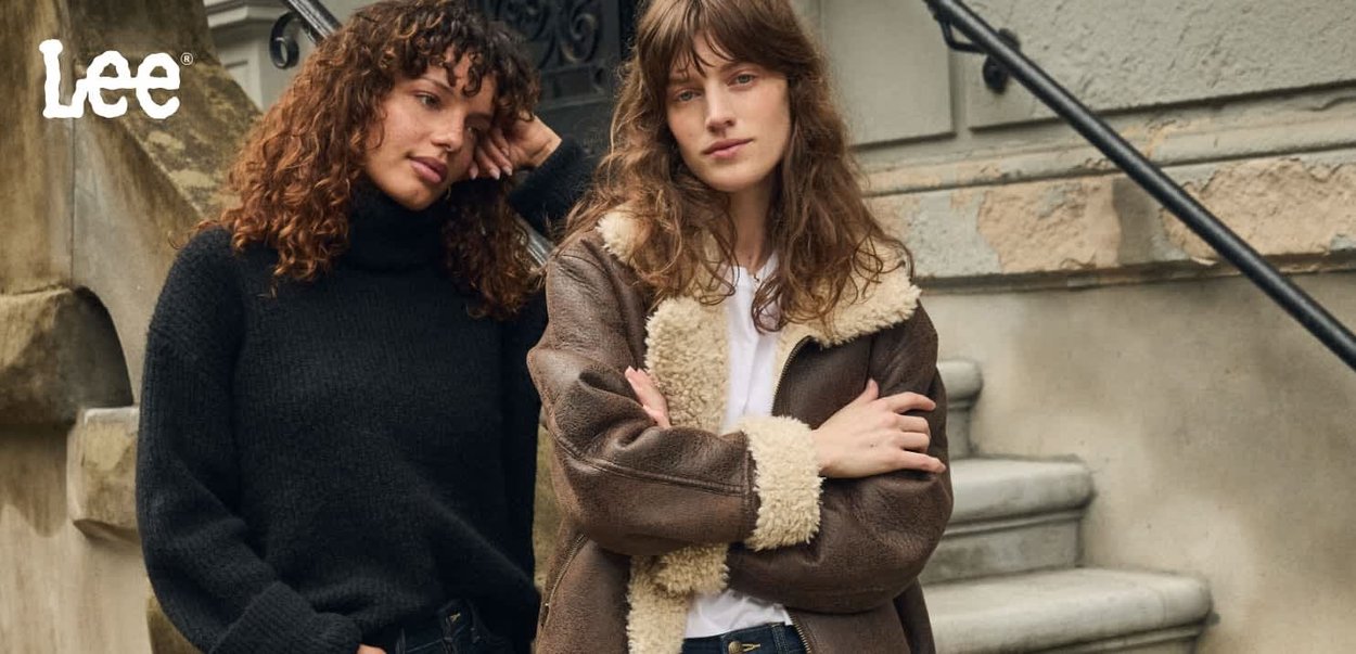 Twee vrouwen poseren buiten. De ene draagt een zwarte gebreide coltrui; de andere een bruine jas met een beige schapenvachtkraag. Trappen en muur op de achtergrond.