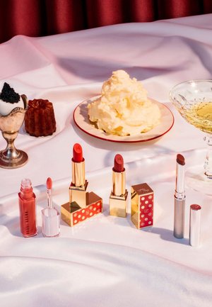 Deux rouges à lèvres rouges avec des boîtiers dorés, dont un dans une boîte décorée, accompagnés d'un gloss à lèvres rose translucide. Une coupe à dessert et une assiette en argent avec de la crème fouettée.