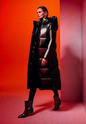 Manteau doudoune noir avec capuche, finition brillante, texture matelassée, associé à des bottes texturées à la cheville. Mis en valeur sur un fond vibrant d'orange et de rose.