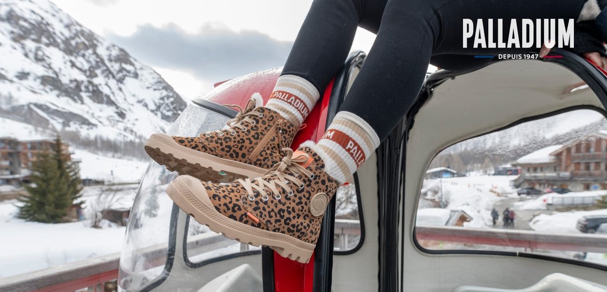 Bottes à motif léopard avec des accents beiges, semelle en caoutchouc texturé et lacets beiges. Portées avec des chaussettes rayées sur fond de montagne enneigée.