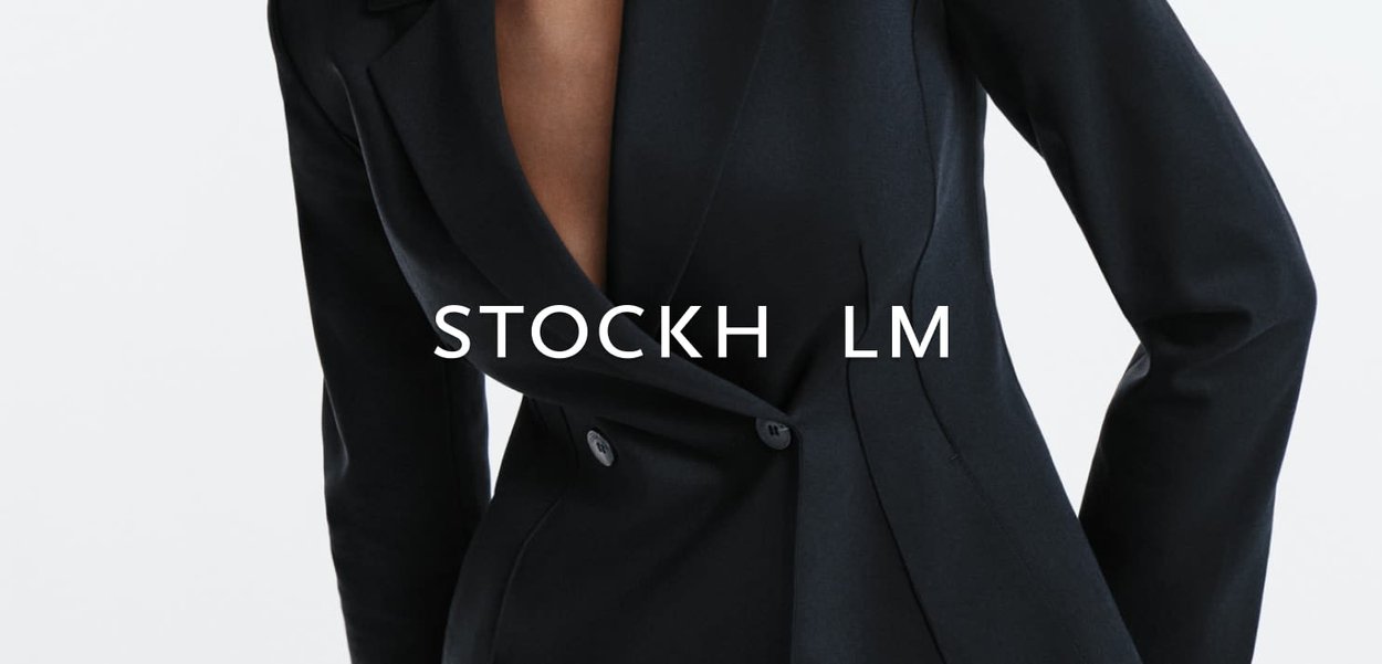 Person trägt ein enganliegendes schwarzes Sakko mit tiefem Ausschnitt und posiert vor einem schlichten weißen Hintergrund.