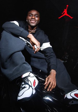 Giovane uomo in tuta bianca e nera seduto con una gamba sollevata, indossa scarpe da ginnastica bianche e rosse, sorride davanti a uno sfondo scuro con il logo Jumpman rosso.