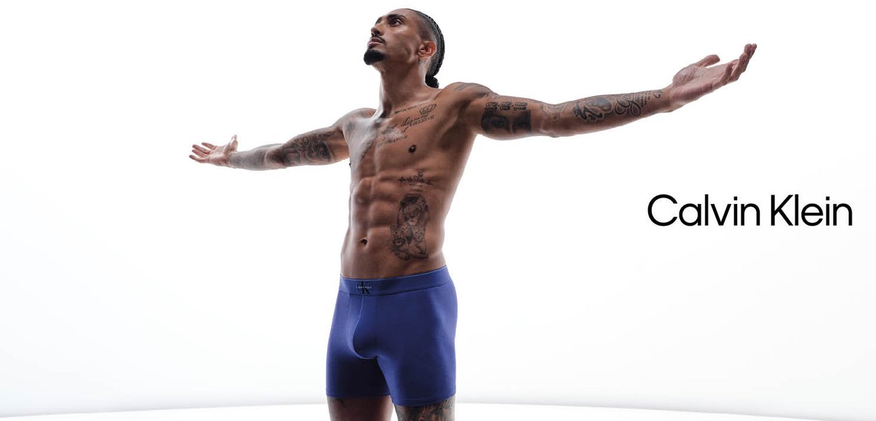 Mężczyzna bez koszulki z tatuażami stoi z rozpostartymi ramionami, ubrany w niebieskie bokserki Calvin Klein na białym tle.