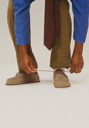 Person trägt olivfarbene Hose, blaues Hemd und braune Krawatte und beugt sich nach vorne, um beige Wildlederschuhe mit weißen Socken auf hellem Hintergrund zu binden.