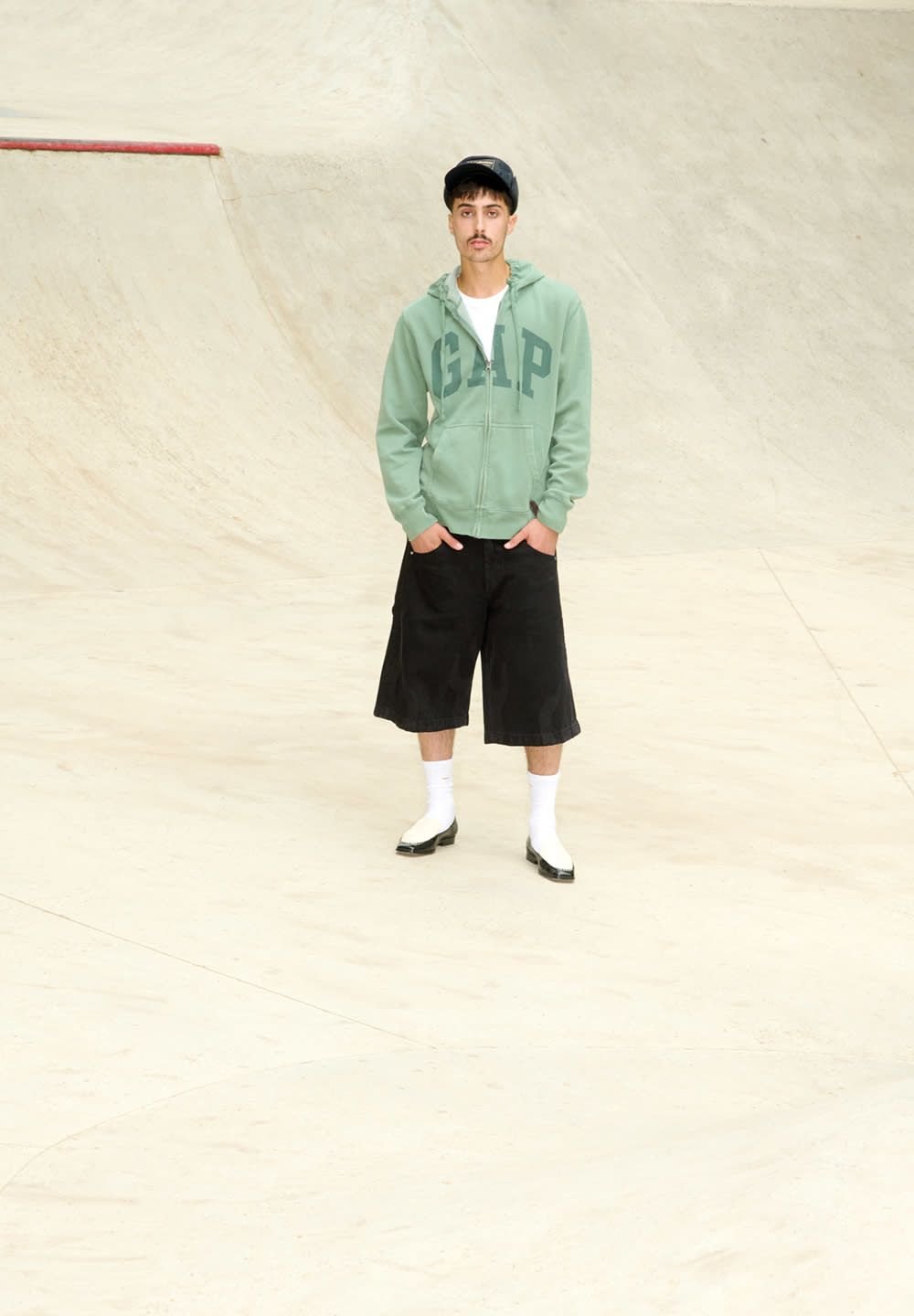 Groene zip-up hoodie met "GAP" logo, zwarte knielange shorts, witte sokken en zwarte schoenen; staande op een glad skateparkoppervlak.