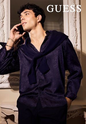 Camisa azul marino con estampado, de mangas largas, con un corte holgado y cierre de botones al frente, combinada con un suéter oscuro sobre los hombros.