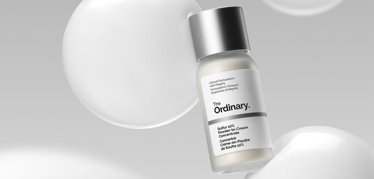 Clear glass bottle with a silver cap, labeled "The Ordinary. Sulfur 10% Powder-to-Cream Concentrate." Two white spherical accents in the background.