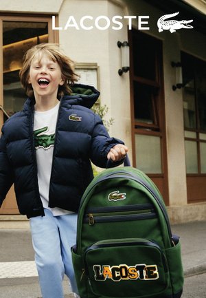 Grüner Lacoste-Rucksack mit gesticktem Logo, Reißverschlussfächern und gepolsterten Trägern. Das Kind trägt eine marineblaue Steppjacke über einem weißen Hemd.