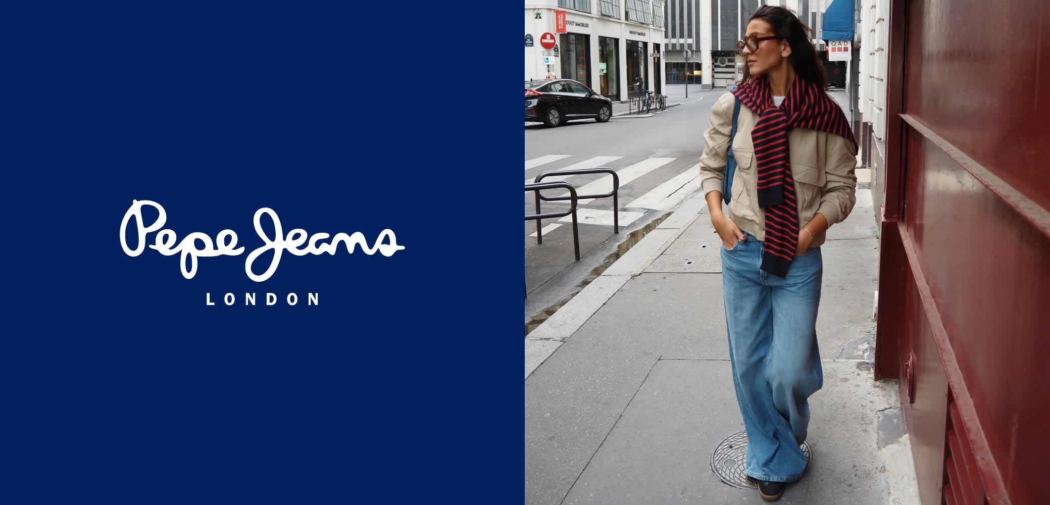 Beige jacket with dual chest pockets, blue wide-leg jeans, and a red-striped scarf worn casually. Background features urban street elements.