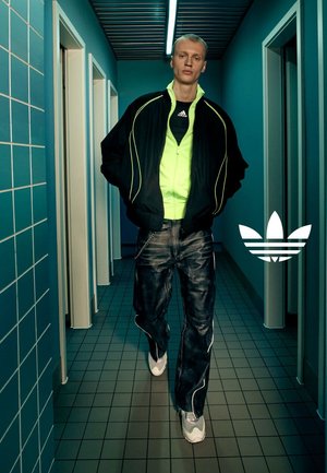 Zwarte en neon groene sportjack, zwarte graphic T-shirt, blauwe denim jeans met witte stiksels en grijze sneakers. Witte Adidas-logo op de vloer.