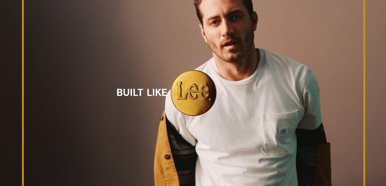 Hvit t-skjorte med korte ermer og lomme, lagd under en brun og svart stripete jakke. Gull sirkulært "Lee" logo tydelig vist.