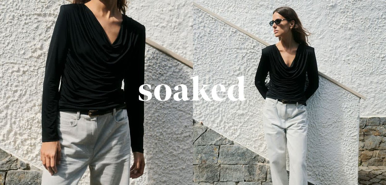 Schwarzes Langarmoberteil mit drapiertem Ausschnitt, kombiniert mit hellen, locker sitzenden Hosen. Im Hintergrund befindet sich eine strukturierte weiße Wand und Treppen.