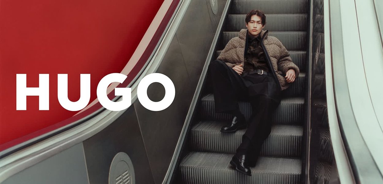 Chaqueta marrón acolchada sobre una camisa negra, pantalones negros y zapatos negros brillantes, sentado en una escalera mecánica con un fondo rojo.