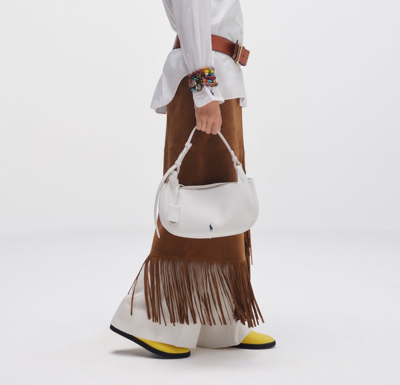 Persoon met een bruine rok met franjes, wit hemd, kleurrijke armbanden, gele schoenen, een witte leren handtas met logo vasthoudend tegen een witte achtergrond.