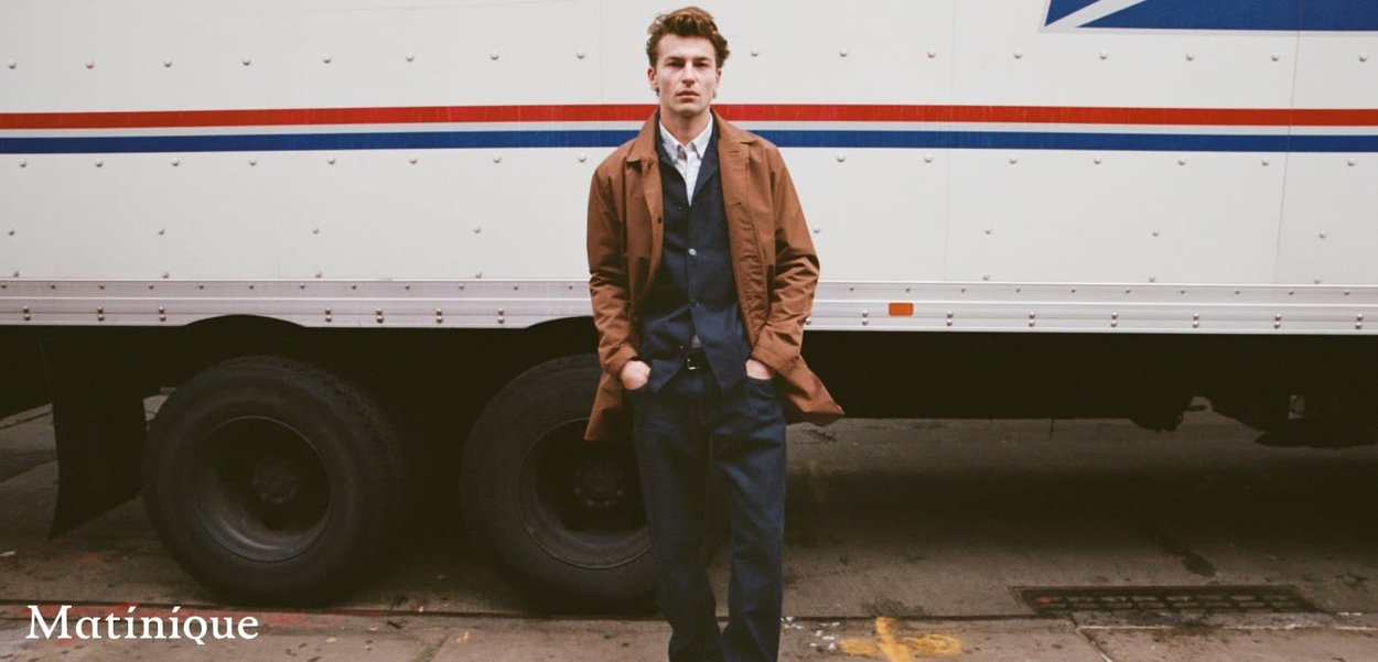 Brauner Mantel über einer dunklen Weste und Hemd, kombiniert mit marineblauen Hosen. Steht vor einem weißen Lastwagen mit roten und blauen Akzenten.