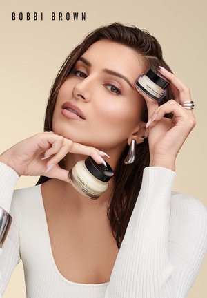 Une femme avec les cheveux mouillés tient deux pots de Bobbi Brown Vitamin Enriched Face Base près de son visage, portant un haut blanc et des boucles d'oreilles en argent.
