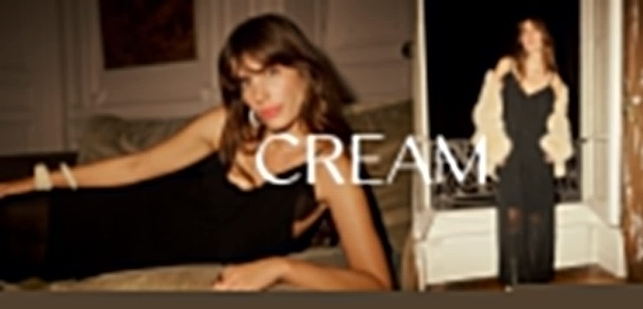 Das schwarze Kleid hat ein tailliertes Design mit dünnen Trägern. Das Model ist in einen cremefarbenen Schal gehüllt und präsentiert sich in eleganten Innenräumen.