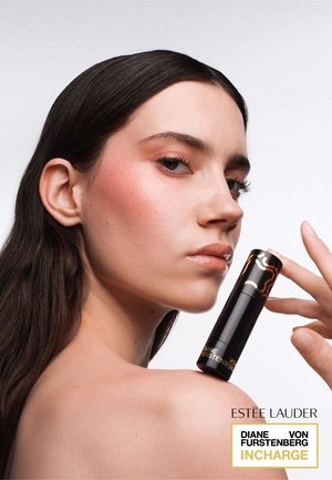 Donna con lunghi capelli scuri tiene vicino alla spalla un rossetto Estee Lauder Diane Von Furstenberg InCharge nero e oro su sfondo bianco.