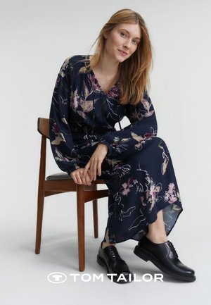 Blumenkleid in Marineblau mit V-Ausschnitt, taillierter Taille und langen Ärmeln. Kombiniert mit schwarzen Schnürschuhen. Sitzt auf einem Holzstuhl.