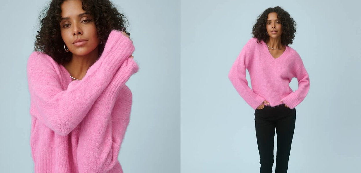 Frau mit lockigem Haar, die einen rosa flauschigen Pullover und schwarze Hose trägt und vor einem hellblauen Hintergrund posiert.