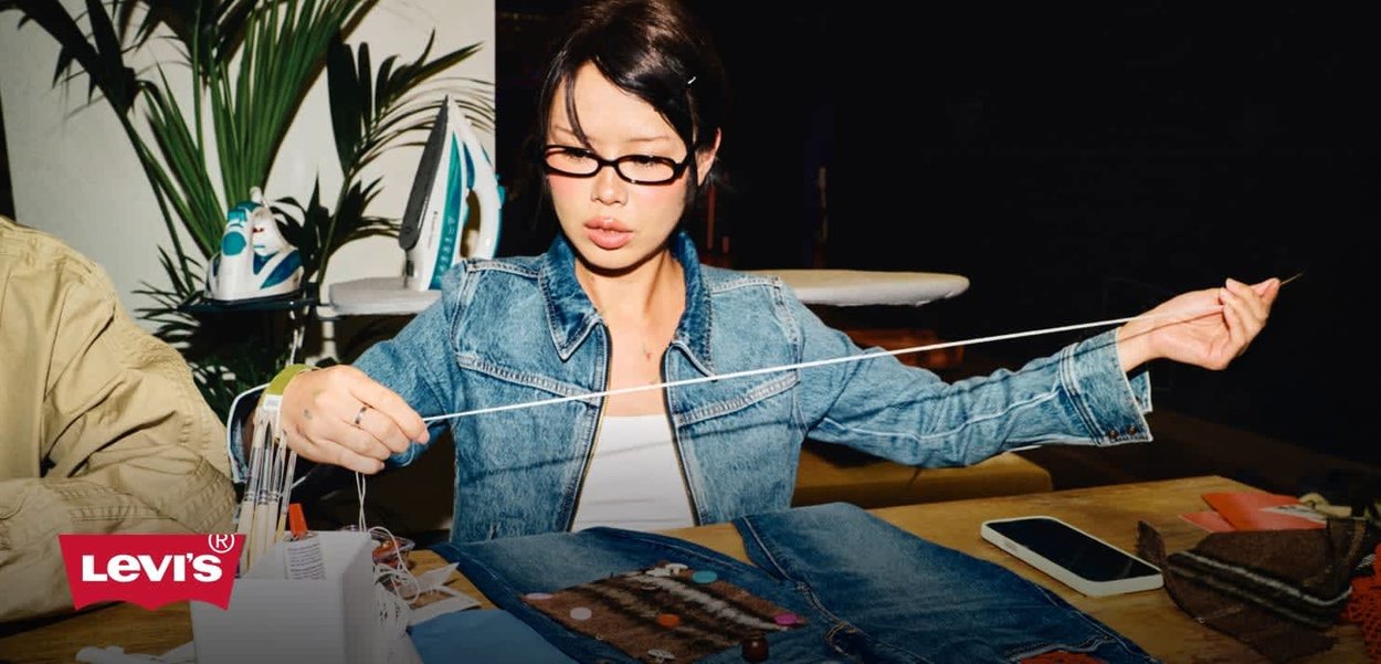 Denim jacket with light wash, worn over a white top. Hands threading a needle with colorful buttons and fabrics on a wooden table. Iron in background.