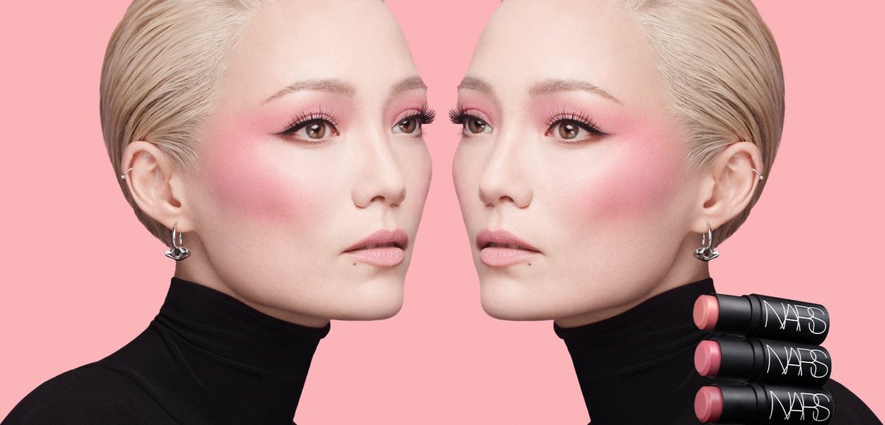 I prodotti labbra NARS esposti accanto a un modello con capelli corti platino, che indossa un rossetto e un trucco occhi audaci in rosa, su uno sfondo rosa tenue.
