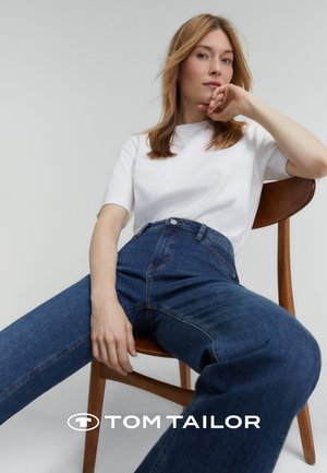 Jeans in denim blu a vita alta con una vestibilità ampia, abbinati a una camicia bianca a maniche corte, seduto su una sedia di legno.